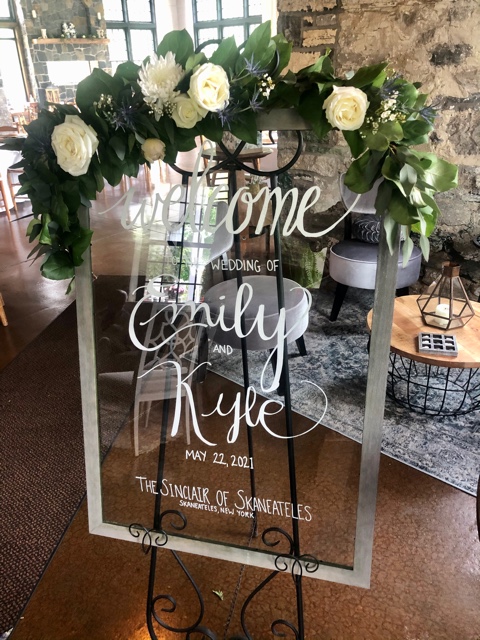 A sign decorate with white flowers that says "Welcome Emily and Kyle"